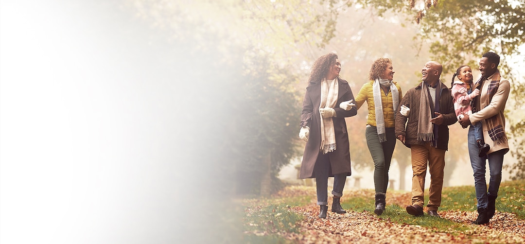Four adults, representing different generations, walk together on a leaf-covered path in a park during autumn, with one carrying a young child.